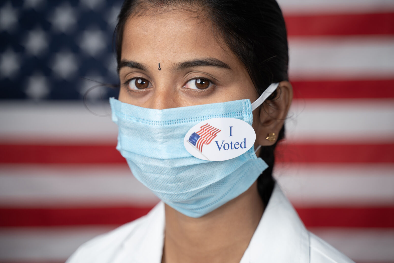 Girl with I voted sticker on medical face mask with US flag as background - concept of US election.